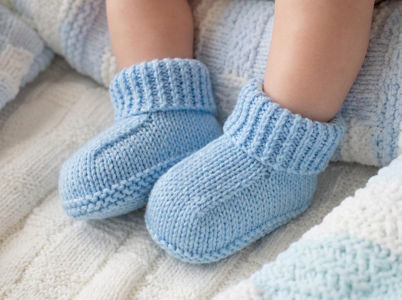 May include: A pair of light blue knitted baby booties. The booties have a ribbed cuff and a simple, classic design. They are shown on a soft, textured blanket, suggesting comfort and warmth.