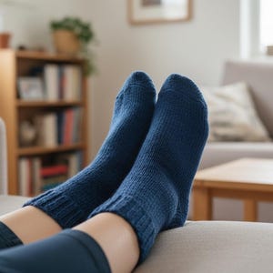 May include: A close-up shot of two navy blue knitted socks. The socks are thick and textured, with a ribbed cuff. The person wearing the socks is relaxing on a light gray couch in a living room setting.