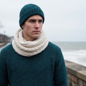Op de afbeelding: Een persoon draagt een teal gebreide beanie, een crèmekleurige gebreide col sjaal en een teal trui. De persoon staat buiten in de buurt van een water. De kledingstukken lijken van wol te zijn.