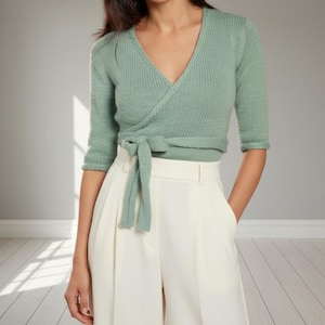 May include: A light green wrap top with a V-neck and three-quarter sleeves, tied at the waist with a matching fabric belt. Paired with cream-colored, high-waisted pants. The outfit is simple and elegant.