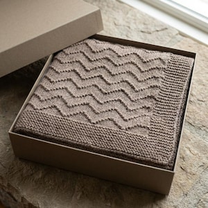 May include: A taupe-colored knitted blanket with a chevron pattern is presented in an open cardboard box. The blanket has a ribbed border and a textured design. The box is also taupe, and the blanket is neatly folded inside, suggesting a gift item.