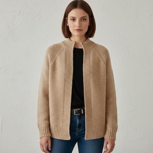 May include: A beige open-front cardigan with a ribbed collar and cuffs. The cardigan is worn over a black top and blue jeans. A gold-coloured belt is visible. The cardigan is made of a soft, textured knit material.