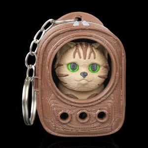 May include: A brown cat keychain with a detailed 3D-printed design. The cat has green eyes and is inside a brown frame with three circular holes. The keychain includes a silver chain and ring.