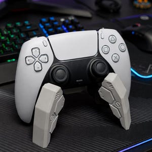 May include: A white and black gaming controller with gray support stands. The controller has a symmetrical design with black joysticks and action buttons. The support stands are gray and angular, providing stability.