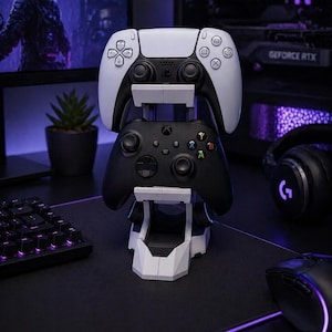 May include: A white and black gaming controller stand holding a white and black PlayStation 5 controller and a black Xbox controller. The stand is white and grey, with a geometric design. The background includes a computer monitor and other gaming accessories.