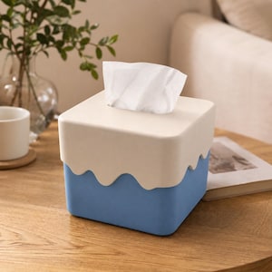 Puede incluir: Caja de pañuelos de dos tonos con una parte superior beige claro y una base azul. La parte superior tiene un diseño de borde ondulado. Los pañuelos blancos sobresalen de la parte superior. La caja está sobre una mesa de madera.