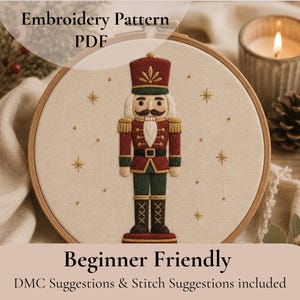 May include: An embroidery pattern PDF featuring a nutcracker design stitched on off-white fabric within a wooden hoop. The nutcracker wears a red and green uniform with gold accents, surrounded by small embroidered gold stars on the fabric.