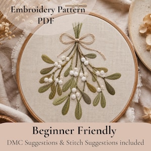 May include: An embroidery pattern PDF featuring a mistletoe design stitched onto light beige fabric within a circular wooden hoop. The mistletoe has green leaves and white berries, tied with a tan bow. Text reads Embroidery Pattern PDF.