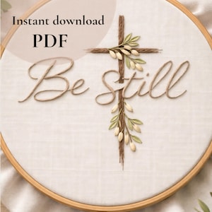 Pode incluir: Um bastidor de bordado com as palavras "Be Still" bordadas em itálico. Uma cruz feita de barbante marrom é adornada com detalhes florais verdes e brancos. O texto "Instant download PDF" é visível no topo.