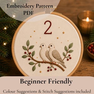 Puede incluir: Un patrón de bordado PDF en un bastidor de madera circular sobre tela color crema. El diseño muestra dos aves blancas posadas en una rama marrón con hojas verdes y bayas rojas, coronadas por el número 2 y pequeñas estrellas doradas.