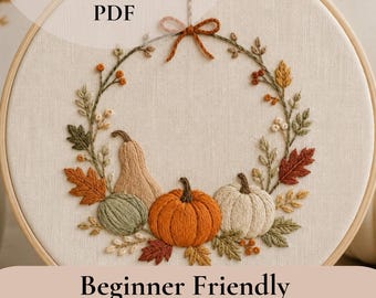 Puede incluir: Un patrón de bordado PDF con un diseño de corona circular sobre tela color crema en un bastidor de madera. La corona incluye calabazas, hojas de otoño y bayas bordadas en naranja, verde y beige. Texto: Embroidery Pattern PDF, Beginner Friendly, DMC Suggestions & Stitch Suggestions included.