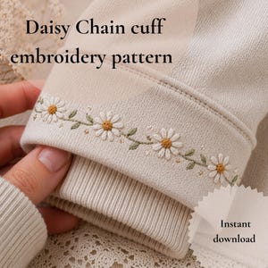 Puede incluir: Primer plano de un puño de sudadera beige con un patrón de bordado de cadena de margaritas. El puño está adornado con margaritas bordadas en blanco y amarillo, con hojas verdes. El texto "Daisy Chain cuff embroidery pattern" es visible.