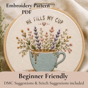 May include: An embroidery pattern PDF featuring a light blue cup overflowing with colorful lavender and wildflower stems on white fabric. The text reads HE FILLS MY CUP with a small heart. The design is held in a circular wooden embroidery hoop.