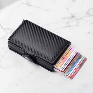 May include: A black card holder with a carbon fiber texture pattern. The rectangular accessory features a metallic frame and a side trigger mechanism that fans out a stack of multi-colored credit and identification cards from the interior compartment.