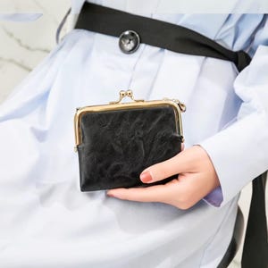 May include: A small black wallet with a textured surface and a gold-toned metal kiss-lock clasp. The rectangular wallet features a smooth, shiny finish on the metal frame and a slightly wrinkled, matte texture on the black material body.
