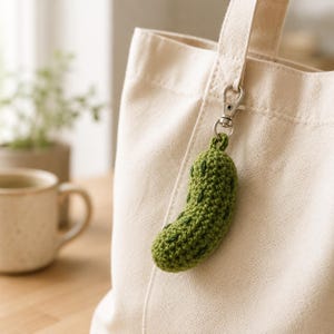 Pode incluir: Um chaveiro de picles verde de crochê com detalhes em verde escuro pendurado em um fecho prateado preso a uma sacola creme. O pingente em forma de picles é feito de fio texturizado.