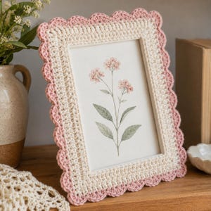 Può includere: Una cornice rettangolare con un design all'uncinetto. La cornice è bianco sporco con un bordo rosa smerlato. Contiene un'immagine di una stampa botanica floreale con fiori rosa e foglie verdi. La cornice è esposta su una superficie di legno.