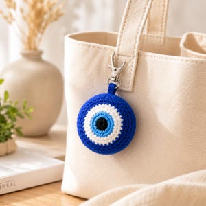 Puede incluir: Un amuleto de crochet azul y blanco con forma de ojo turco con un cierre plateado, sujeto a un bolso tote de lona beige. El amuleto presenta un centro negro, rodeado de círculos concéntricos azules y blancos.