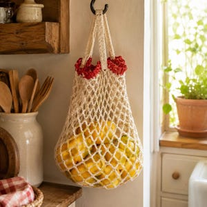 May include: A cream-colored, crocheted net bag filled with bright yellow lemons, hanging from a black hook. The bag has red decorative trim at the top. The bag is hanging in a kitchen setting.