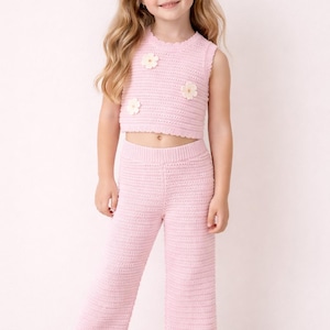 Puede incluir: Un conjunto de dos piezas de crochet rosa claro para niña. El conjunto incluye un top corto sin mangas adornado con tres aplicaciones de flores color crema y pantalones de pernera ancha a juego. La niña también lleva zapatos blancos.