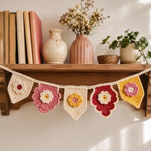 Peut inclure: Une guirlande décorative au crochet avec des motifs floraux aux couleurs crème, rose, rouge et jaune. La guirlande est suspendue sur une étagère en bois avec des livres, des vases et des plantes.
