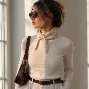 May include: A woman wearing a cream-colored turtleneck shirt, light-colored pants, and a brown leather belt. She is also wearing a small, brown shoulder bag, sunglasses, and a knitted scarf. The outfit is simple and stylish.