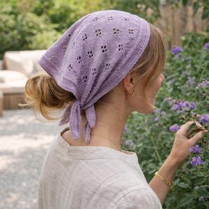 May include: A light purple knitted headscarf with a delicate openwork pattern. The scarf is tied at the back and worn on the head. The headscarf is made of a soft, lightweight material.