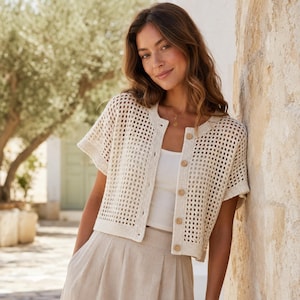 Puede incluir: Rebeca de crochet color crema con mangas cortas y botones de madera. El diseño de tejido abierto revela una camiseta sin mangas blanca debajo. La rebeca se combina con pantalones beige, creando un atuendo neutro y veraniego.