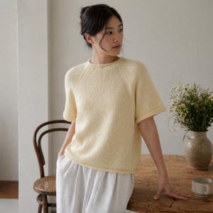 Puede incluir: Un suéter de punto amarillo pálido de manga corta con cuello redondo. El suéter tiene un aspecto ligeramente texturizado y un ajuste relajado. Las mangas están dobladas. La modelo lleva el suéter con pantalones blancos. El suéter es de color claro y neutro.