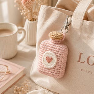 Puede incluir: Un llavero de botella de perfume rosa tejido a ganchillo con una tapa beige y un círculo blanco con un corazón rosa. El llavero está sujeto a un bolso tote beige con la palabra "LOVE" impresa. Una taza de café y un libro están en el fondo.