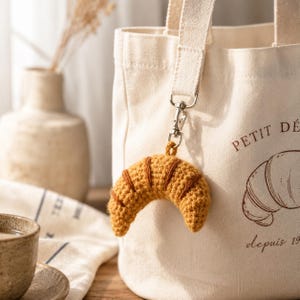 May include: A crocheted croissant keychain in shades of brown and tan hangs from a canvas tote bag. The bag has the words "PETIT DÉ" and "depuis 19" printed on it. A cup of coffee and a vase are in the background.