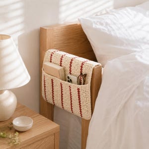 May include: A cream-colored, crocheted bedside organizer with vertical red stripes is attached to a wooden headboard. The organizer holds a book, a smartphone, and eyeglasses. A white bed and a wooden nightstand with a lamp are in the background.