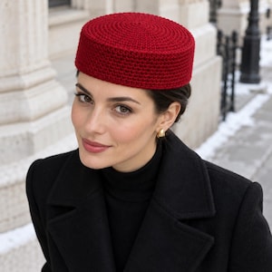 May include: A deep red, cylindrical pillbox hat crafted with a dense, textured crochet stitch pattern. The hat features a flat top and straight, structured sides, providing a solid, uniform appearance in a vibrant, saturated crimson hue.