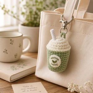Könnte beinhalten: Ein gehäkelter Matcha-Latte-Schlüsselanhänger mit weißem Oberteil und grünem Boden, befestigt an einer beigen Tragetasche. Der Schlüsselanhänger hat einen weißen Strohhalm und ein Etikett mit der Aufschrift "Matcha Latte". Eine Tasse und ein Notizbuch sind im Hintergrund.