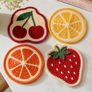 Puede incluir: Cuatro posavasos de ganchillo hechos a mano con forma de fruta. Los posavasos incluyen una cereza, un limón, una rodaja de naranja y una fresa. El posavasos de cereza es rojo con un tallo y una hoja verdes. El limón es amarillo, la naranja es naranja y la fresa es roja.