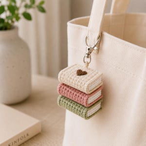 May include: A stack of three crocheted books in cream, pink, and green, attached to a silver keychain. The top book has a brown heart. The keychain is hanging from a white tote bag.