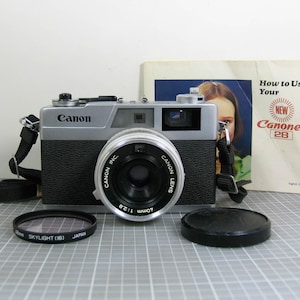 May include: A silver and black Canon Canonet 28 camera with a 40mm lens, lens cap, and Skylight (1B) filter. The camera has a black strap and is accompanied by a manual titled "How to Use Your NEW Canonet 28."