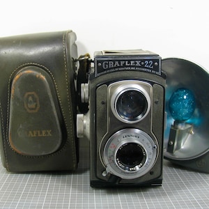 May include: A vintage Graflex 22 twin-lens reflex camera with a Century lens, a metallic flash reflector with a blue bulb, and a dark green leather carrying case. The camera body features the text GRAFLEX 22 and MADE IN USA BY GRAFLEX INC ROCHESTER NY.