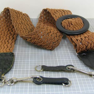 May include: A brown woven belt with a black buckle and leather accents. The belt has a braided design and includes two detachable black leather straps with metal clasps. The belt is approximately 40 inches long.
