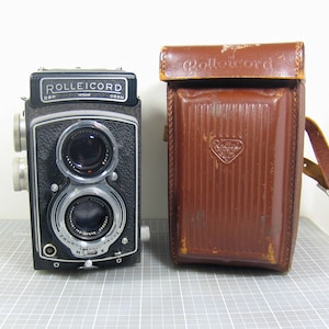 May include: A vintage Rolleicord camera with a leather case. The camera is black with silver accents and two lenses. The leather case is brown with a shoulder strap and the word "Rolleicord" embossed on the top.