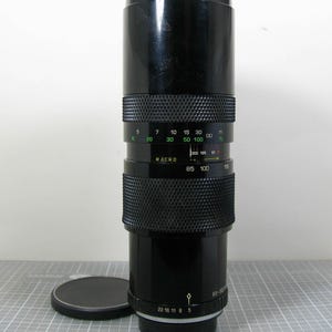 May include: A black camera lens with a textured grip and a lens cap. The lens has markings for focal lengths and aperture settings. The lens is approximately 12 inches long.