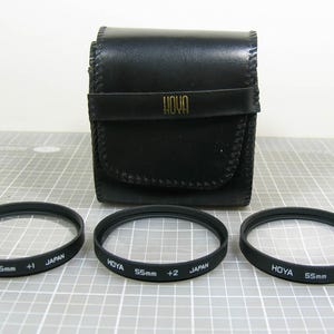 May include: A black leather case with the word "HOYA" in gold lettering, and three black 55mm camera lens filters. The filters are labeled with "HOYA", "55mm", and "+1", "+2", and "+4" and "JAPAN".