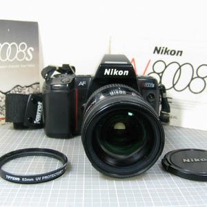 May include: A black Nikon N8008s 35mm film camera with an attached lens, a black camera strap, a Tiffen 62mm UV protector filter, a black Nikon lens cap, and two Nikon N8008s instruction manuals with white covers and black text.