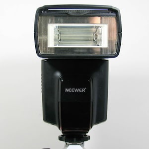 May include: A black camera flash with the brand name "NEEWER" printed on the front. The flash has a rectangular head with a clear, ridged light panel. The flash is mounted on a camera.