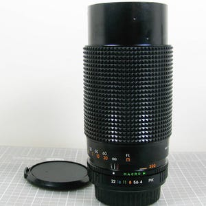May include: A black camera lens with a textured rubber focus ring, featuring orange distance markings in feet and meters, a green macro setting, and white aperture numbers ranging from 4 to 22. A separate black circular lens cap sits nearby.