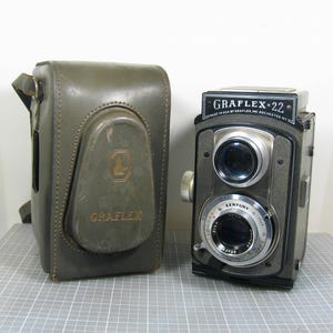 May include: A vintage Graflex 22 twin-lens reflex camera with a black body and silver lens housing, featuring the text GRAFLEX 22 MADE IN USA BY GRAFLEX INC ROCHESTER NY. Beside it sits a matching olive green leather carrying case with GRAFLEX embossed.