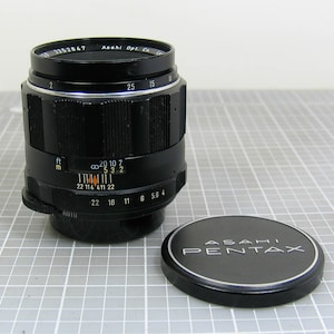 May include: A black Asahi Pentax camera lens with a matching lens cap. The lens has various measurement markings and a focus ring. The lens cap is black with the brand name in white lettering. The lens is approximately 3 inches tall.