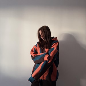 May include: A person wearing a striped hoodie in shades of orange, teal, and brown, paired with black pants. The hoodie features a drawstring and a relaxed fit. The background is a plain, light-colored wall.