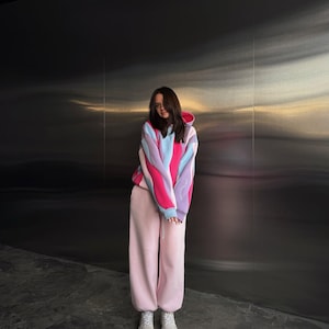 May include: A person wearing a color-block hoodie in pink, blue, and purple, paired with light pink sweatpants and white sneakers. The hoodie has a drawstring hood and long sleeves. The outfit is casual and comfortable.