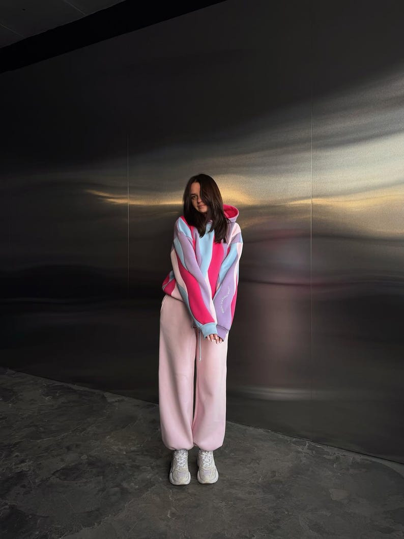 May include: A person wearing a color-block hoodie in pink, blue, and purple, paired with light pink sweatpants and white sneakers. The hoodie has a drawstring hood and long sleeves. The outfit is casual and comfortable.
